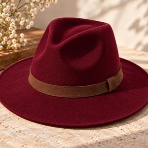 Könnte beinhalten: Ein tiefroter Fedora-Hut mit breiter Krempe und strukturierter Krone. Der Hut ist mit einem glatten, braunen Kunstlederband akzentuiert, das um die Basis der Krone verläuft und mit einer kleinen, passenden braunen Schlaufe an der Seite befestigt ist.