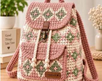 May include: A crochet backpack featuring a diamond pattern in dusty pink, cream, and sage green. The bag has a top handle, two shoulder straps, a front pocket, and a brown leather-like strap closure with a brass-toned metal clasp and a small tag.