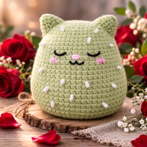 May include: A light green crocheted cat-shaped plush toy with white accents, closed eyes, and pink cheeks, resting on a wooden slice. Red roses and scattered petals surround the toy, creating a romantic and handcrafted aesthetic.