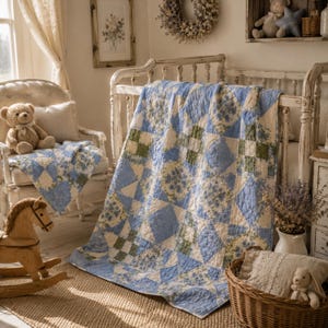 May include: A blue, white, and green patchwork quilt draped over a white crib. The quilt features floral and geometric patterns. A matching quilt is on a white chair with a teddy bear. A wooden rocking horse is in the foreground.