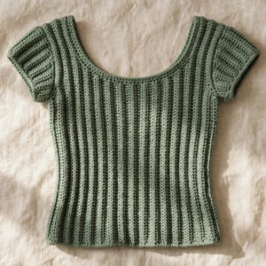 Puede incluir: Una blusa de manga corta en color verde salvia confeccionada con un patrón de ganchillo de canalé vertical. La prenda presenta un amplio escote redondo y mangas cortas, mostrando una construcción de punto texturizado uniforme.