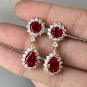 Puede incluir: Un par de pendientes colgantes dorados con grandes gemas rojas vibrantes en forma de lágrima, rodeadas de piedras blancas más pequeñas y brillantes. Los pendientes tienen un diseño floral en la parte superior e inferior.