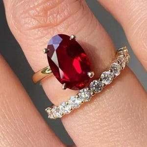 May include: A gold ring with a large oval-cut ruby gemstone and a smaller band with multiple small, clear gemstones. The ruby is a deep red color, and the smaller band is set with clear, sparkling stones.