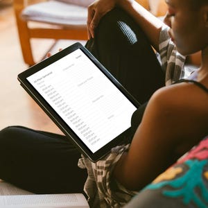 Puede incluir: Una tableta negra que muestra un horario es sostenida por una persona sentada en un sofá con un libro y un cojín estampado. La pantalla de la tableta muestra una lista de horas y eventos, de 06:00 a 23:00.