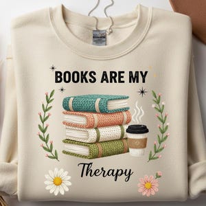 Puede incluir: Sudadera color crema con el texto "BOOKS ARE MY Therapy". El gráfico presenta una pila de libros coloridos, una taza de café y detalles florales. Los libros son turquesa, naranja, blanco y verde.