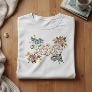 Può includere: T-shirt bianco girocollo con design "XOXO" ricamato. Il design presenta le lettere "XOXO" in un carattere strutturato bianco sporco, decorate con spruzzi colorati a forma di cuore e circondate da composizioni floreali con rose blu, rosa e gialle.