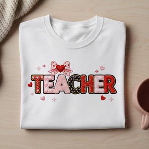 May include: White t-shirt with the word "TEACHER" in a decorative font. The letters are red, pink, and black, with a bow and hearts above the word. The shirt is folded on a light wood surface.