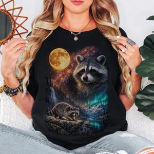 Puede incluir: Una camiseta negra con un estampado gráfico de la cara de un mapache y un mapache más pequeño sobre un acantilado rocoso. La escena incluye una luna llena, un cielo estrellado, una cascada, montañas y auroras boreales sobre un lago.