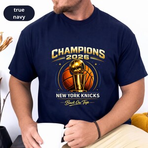 May include: A true navy short-sleeve t-shirt featuring a graphic with the text CHAMPIONS 2026, NEW YORK KNICKS, and Back On Top. The design includes a gold basketball trophy centred in front of a large orange basketball with gold accents.