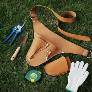 Op de afbeelding: Een verzameling tuingereedschap en accessoires, waaronder een bruine leren gereedschapsriem, een blauw-zilveren snoeischaar, een kleine troffel, een groen-gele rolmaat, een bruine leren holster en witte tuinhandschoenen.