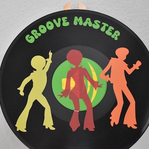 Puede incluir: Un disco de vinilo negro con la inscripción "GROOVE MASTER" en verde. Siluetas de tres figuras bailando en amarillo, rojo y naranja están en el disco. Un círculo verde está en el centro.