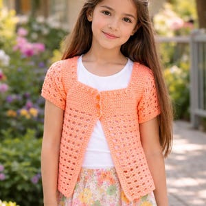 May include: A peach-colored, short-sleeved crochet cardigan featuring a square neckline and three matching buttons down the front. The garment is crafted with a solid stitch pattern at the top and a decorative openwork stitch design throughout.