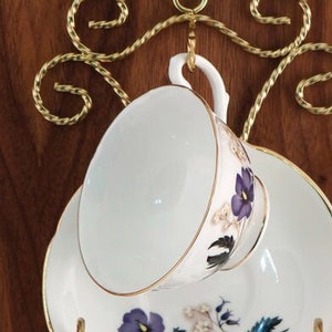 May include: A gold metal teacup and saucer holder with a decorative swirl design. The holder is attached to a wall and has a teacup and saucer hanging from it.