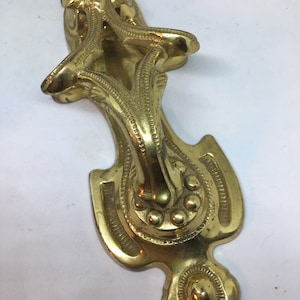 May include: A gold-toned metal door knocker with an ornate design. The knocker has a floral motif and a ring-shaped handle.