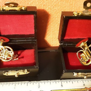 May include: Two miniature gold musical instruments, a French horn and a tuba, are displayed in their individual black velvet-lined cases with brass clasps.