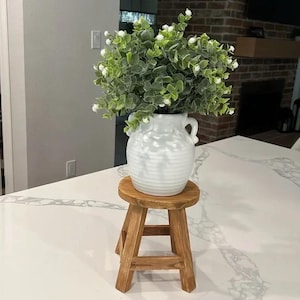 May include: A white ceramic vase with two handles, filled with green and white faux eucalyptus, sits on a small, round, wooden stool. The stool has a natural wood finish and a three-legged design. The vase has a ribbed texture.