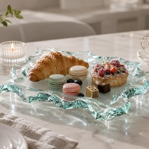 Könnte beinhalten: Ein Serviertablett aus klarem Glas mit strukturiertem, gewelltem Rand, auf dem ein goldbraunes Croissant, drei bunte Macarons in Hellgrün, Rosa und Weiß, eine kleine Obsttorte mit Himbeeren und zwei kleine Pralinen liegen.
