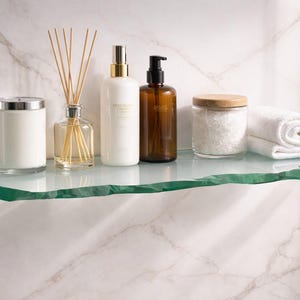 May include: A clear glass shelf with a natural edge holds bathroom accessories. Items include a candle, reed diffuser, lotion bottles, a jar of bath salts, and a rolled white towel. The shelf is mounted on a marble-patterned wall.