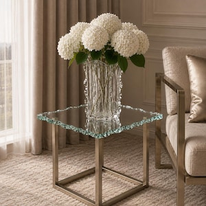Puede incluir: Una mesa auxiliar cuadrada con una superficie de cristal transparente de borde ondulado y texturizado sobre una estructura metálica. Un florero de cristal transparente con estrías verticales contiene un denso ramo de hortensias blancas con tallos verdes.