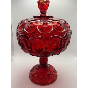 Puede incluir: Un cuenco de cristal rojo intenso con tapa a juego, que presenta un patrón de lunas y estrellas en el cuerpo, la tapa y la base. El recipiente tiene una forma redondeada, un borde acampanado y un remate decorativo en la parte superior.