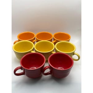 Op de afbeelding: Een set van acht keramische koffiemokken in verschillende kleuren. De mokken zijn in twee rijen gerangschikt, met kleuren als oranje, geel en rood. Elke mok heeft een afgeronde vorm met een oor.