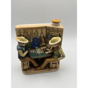 May include: A ceramic flask featuring a poker game scene. The flask is tan with a detailed relief of figures around a table. The figures are wearing hats and vests. The flask has a cork stopper.