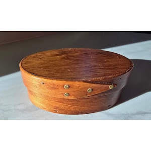Puede incluir: Una caja de madera de forma ovalada con tapa, que presenta una construcción de madera curvada asegurada por una banda superpuesta puntiaguda. La caja está hecha de madera de color marrón medio con veta visible y sujeta con tres pequeños pasadores metálicos.