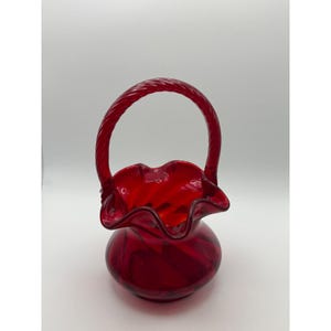 May include: A ruby red glass basket with a twisted handle. The basket has a ruffled rim and a round base. The glass has a marbled appearance. The basket is a decorative item.