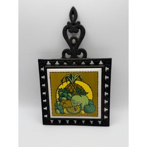 May include: Black cast iron trivet with a square tile featuring a fruit design. The tile has a yellow background with a pineapple, grapes, and other fruits. The trivet has a decorative handle.