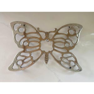 Puede incluir: Un salvamanteles de metal plateado con forma de mariposa. El salvamanteles tiene un diseño recortado con formas redondeadas y ovaladas en las alas y el cuerpo. Está diseñado para proteger las superficies de los utensilios de cocina calientes.