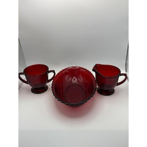 May include: A set of three ruby red glass items comprising a sugar bowl with two handles, a creamer jug with a handle and spout, and a scalloped edge bowl. Each piece features a pedestal base and a smooth, translucent red finish throughout.