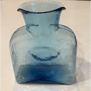 Pode incluir: Uma jarra de vidro azul translúcido com corpo quadrado e borda larga e alargada. O vidro apresenta uma superfície texturizada e ondulada, criando um efeito de reflexo de luz. A base é ligeiramente recuada e a cor torna-se mais profunda.