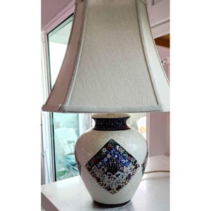 Könnte beinhalten: Dekorative Tischlampe mit cremefarbenem Lampenschirm und weißem Keramikfuß. Der Sockel hat ein rautenförmiges Blumendesign in Blau-, Rot- und Grüntönen. Die Lampe hat einen dunkelbraunen Rand und einen cremefarbenen Lampenschirm.