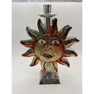 Könnte beinhalten: Ein Kerzenhalter aus Metall mit einem zentralen Sonnengesicht-Design und strahlenden Strahlen. Die Sonne hat eine metallische Oberfläche mit roten und orangefarbenen Akzenten, einem lächelnden Ausdruck und definierten Augen auf einem Sockel.