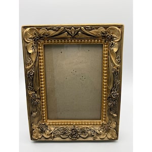 Peut inclure: Un cadre photo rectangulaire doré avec des détails floraux ornés. Le cadre présente des roses et d'autres éléments décoratifs sur les bords et une bordure intérieure perlée. Le cadre est conçu pour contenir une photographie ou une œuvre d'art.