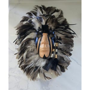 May include: A decorative mask with a face-shaped center, surrounded by gray and black feathers. The face is painted in a light skin tone with black and blue accents. Black ribbons and beaded strands add detail.