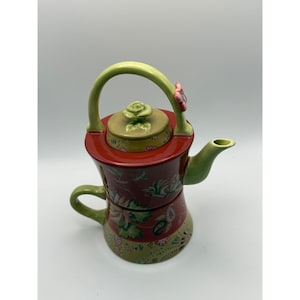 May include: A decorative ceramic teapot with a matching teacup. The teapot is red with floral designs, a green handle, and a green spout. The lid is tan with a rose-shaped knob. The teacup is stacked beneath the teapot.
