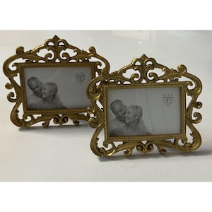 Può includere: Due cornici dorate ornate con un'apertura rettangolare per foto. Le cornici presentano un design a volute dettagliato. Ogni cornice contiene una foto in bianco e nero di una coppia. Il marchio "ELSA" è visibile.