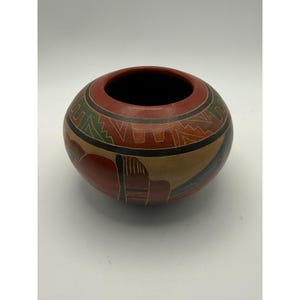 May include: A round, low-profile ceramic bowl with a smooth, matte finish. The exterior features a terracotta red base color decorated with geometric patterns in black, tan, and green, accented by thin, incised white lines throughout the design.