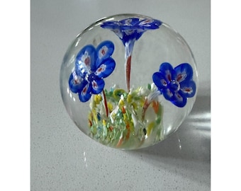 May include: A clear glass paperweight shaped as a sphere, containing three blue flowers with white centers and orange stems, set above a base of multicolored glass fragments in shades of green, yellow, and white.