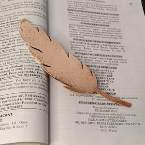 May include: A copper-colored metal feather bookmark rests on an open book. The bookmark has a detailed, textured surface and a stylized feather shape. The book's pages are filled with small text.