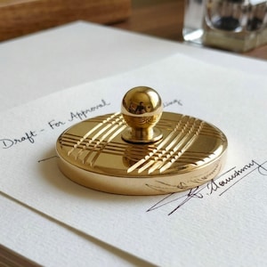 May include: A gold-colored desk stamp with a round top and a small, spherical handle. The stamp has a plaid-like pattern etched into its surface. The words "Draft - For Approval" are written in cursive on the paper beneath the stamp.