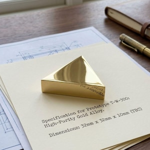 May include: A polished gold alloy triangular object, 32mm x 32mm x 10mm, sits on a cream paper with text. The text reads "Specification for Prototype T-W-300: High-Purity Gold Alloy. Dimensions: 32mm x 32mm x 10mm (TBC)". A pen and notebook are in the background.