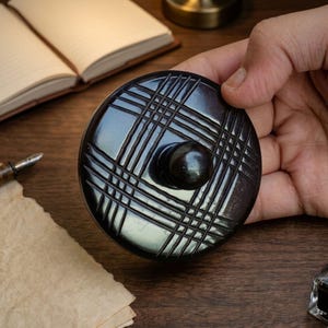 May include: A dark brown, round inkwell lid with a grid pattern and a central knob. The lid is held by a hand, with a blurred background of a desk, notebook, and pen.