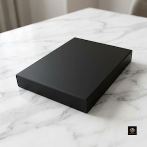 Op de afbeelding: Een rechthoekige, effen zwarte doos met een gladde, matte afwerking. Het object heeft scherpe, strakke randen en een plat bovenoppervlak, wat zorgt voor een eenvoudige, uniforme uitstraling. De doos is in een hoek geplaatst om de zijden te tonen.
