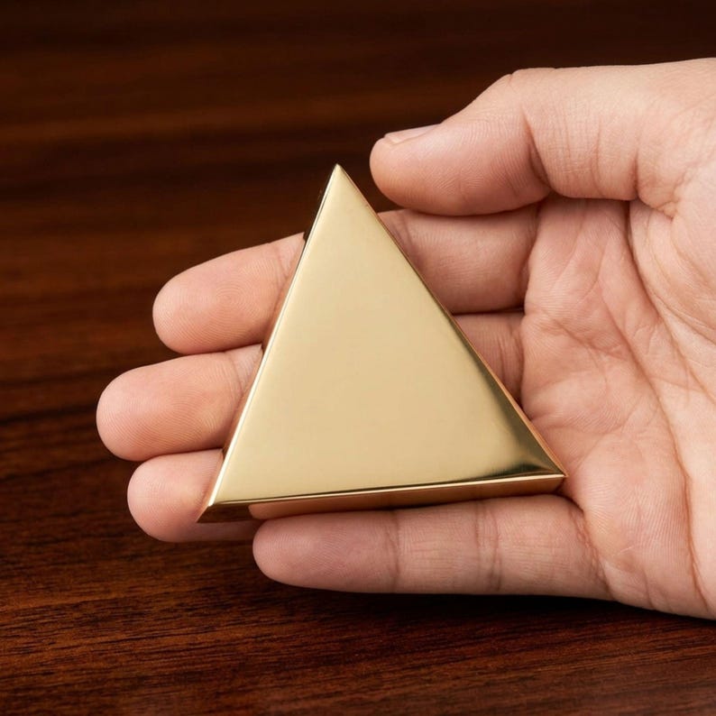 K&ouml;nnte beinhalten: Ein goldfarbenes, dreieckiges Objekt mit polierter Oberfl&auml;che. Das Objekt wird in einer Hand gehalten, wodurch seine geometrische Form und sein metallischer Glanz zur Geltung kommen. Der Hintergrund ist eine dunkle Holzoberfl&auml;che.