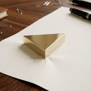 K&ouml;nnte beinhalten: Ein goldfarbener, dreieckiger Briefbeschwerer liegt auf einem Blatt cremefarbenem Papier. Der Briefbeschwerer hat ein solides, geometrisches Design. Weitere B&uuml;roartikel, darunter ein Stift und B&uuml;roklammern, sind im Hintergrund zu sehen.