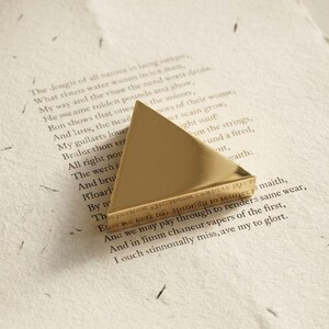 K&ouml;nnte beinhalten: Ein goldfarbener, dreieckiger Briefbeschwerer liegt auf einer Seite mit gedrucktem Text. Der Briefbeschwerer hat eine polierte, reflektierende Oberfl&auml;che. Der Text auf der Seite ist in einer Schriftart geschrieben.