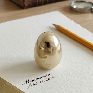 May include: A polished, golden egg-shaped ornament reflecting its surroundings, placed on white paper. The paper bears the handwritten inscription "Memoranda Sept. 15, 2024". A pencil and a magnifying glass are also present.