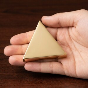K&ouml;nnte beinhalten: Ein goldfarbenes, dreieckiges Objekt mit flacher Basis und glatter, reflektierender Oberfl&auml;che. Der pyramidenf&ouml;rmige Gegenstand wird in einer offenen Hand gehalten, wodurch seine geometrische Form und metallische Oberfl&auml;che zur Geltung kommen. Die Kanten des Objekts sind klar definiert.